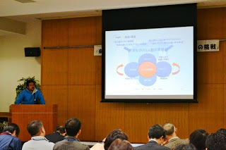 青いジャンパーを着た発表者が演台に立ち、円形図が表示されたスライドを使って説明している様子を聴衆が聞いている写真