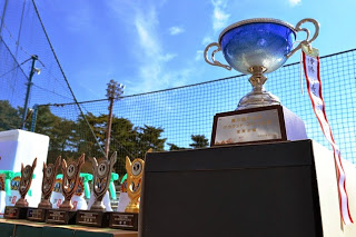 青空を背景に、並べられたカラフルなトロフィーと、左側に大きな優勝カップが置かれている様子の写真