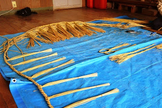 青いシートの上に置かれたしめ縄の製作途中の状態が写っており、手前には長く伸ばした藁が等間隔に並べられている写真