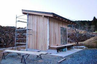 完成に近づいた木の外壁の小屋が足場や作業台の周囲にあり、夕方の光の中にたたずんでいる写真