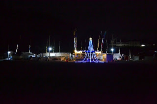 青いLEDの光でクリスマスツリーの形をしたイルミネーションが中央に立ち、その周囲に仮設店舗や旗が並んでいる夜の会場全体を遠くから撮影した写真