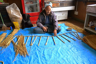 頭に手ぬぐいを巻いた男性が青いシートの上に座り、藁を等間隔に並べ、しめ縄の形を作り始めている様子の写真