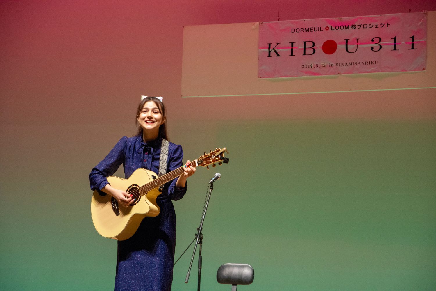 背景はピンクと緑のグラデーションで彩られ、上部には横断幕が掲げられ、マイクスタンドと椅子が置かれたステージ上で、青いワンピースを着て頭に猫耳をつけたセレイナ・アンさんがアコースティックギターを演奏している写真