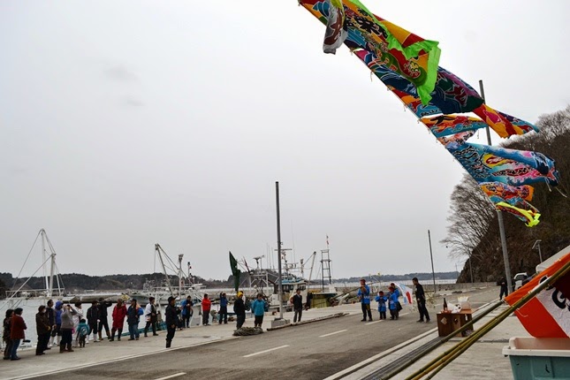 大漁旗が風になびく港で、人々が海沿いに並んでいる写真