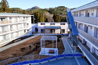 周囲に山や木々が見える3階建ての集合住宅に囲まれた中庭で、作業員が整地をしている新しい団地の写真