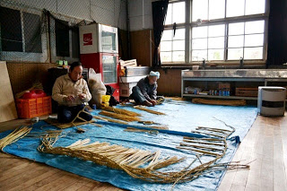 木の床に敷かれた青いシートの上で、2人の男性が藁を使って手作業でしめ縄を編んでいる様子の写真
