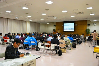 スクリーンが設置された会議室で、多くの参加者が椅子に座って前方の発表を静かに聞いている講演会の全体風景の写真