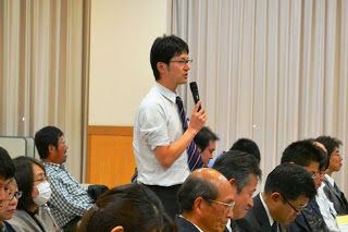 聴衆の中から白いシャツとネクタイ姿の男性が立ち上がり、マイクを使って質問している様子を写した写真