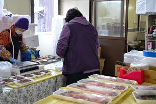 魚介類のパックが並ぶ店内で女性客が商品を購入し、販売員が対応している様子の写真