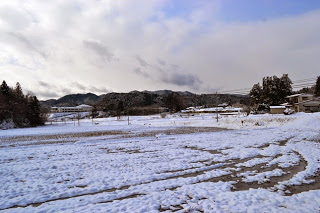雪が一面に積もった田んぼと、遠くに見える山や家々が広がる冬の風景の写真