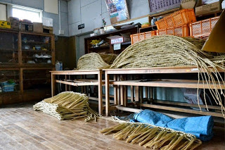 木の机の上に完成したしめ縄が重ねて置かれ、その下に材料の藁束が積まれている室内の写真