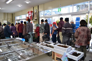 海産物などの商品がテーブルやケースに並べられている店内で、多くの買い物客がレジに並んでいる様子の写真