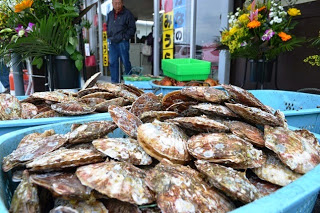 背景に祝い花が飾られている店の外に、青いかごに入った大量のホタテ貝が積まれている写真