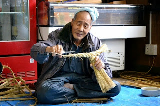 年配の男性が笑顔で右手にハサミを持ち、編んだ藁を手にしながらしめ縄の仕上げ作業をしている写真