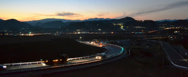 カーブした道路に車やトラックが走っており、夕暮れの山並みと町の灯りが広がっている風景の写真