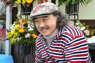 店の外で笑顔を浮かべる3色のストライプの服を着た男性をアップで撮影した写真