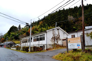 木々が立ち並ぶ山あいの道路沿いに、「さんさん館」と書かれた看板が駐車場の入り口に立っており、奥に白い木造校舎が建っている写真