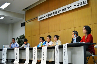 横断幕に「来たるべき将来を見据えた地域づくりへの挑戦」と書かれた前方に、長机に貼られた名前の書かれた札の前に座った7人の登壇者が並んでいる写真