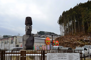 背景に仮設店舗の建物や花輪が見え、手前には大きなモアイ像が立っている写真