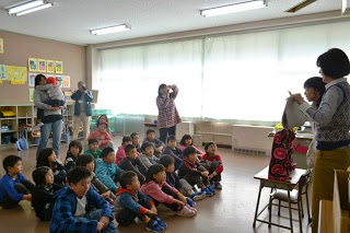 子どもたちが教室の床に座って先生の話を静かに聞いており、関係者がその様子を後ろからカメラで撮影している教室内の様子の写真