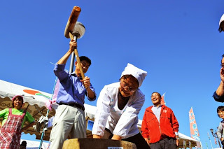 周囲には笑顔の見物人が見守るなか、男性が大きな木槌で餅をつき、女性が合いの手を入れている様子の写真