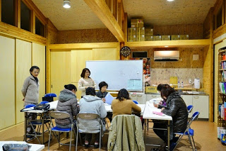 木造の温かみのある室内のホワイトボードの前には講師が立っており、長机を囲んで座った参加者たちが日本語学習をしている写真