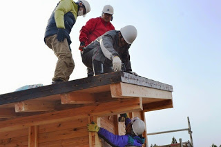 ヘルメットを被った作業員が壁の角を仕上げており、小屋の屋根の上ではヘルメットを被った三人の作業員が話し合いながら作業を進めている写真