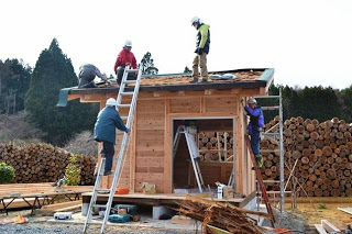 木造の小屋の屋根や壁で、ヘルメットを被った作業員たちが、ハシゴや足場を使って大工仕事をしている写真