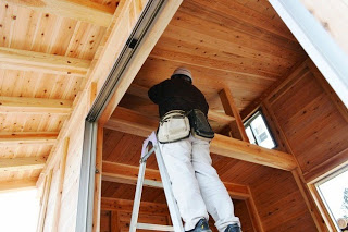 小屋の内部で職人が脚立に登り、天井の梁に木材を組み付けている作業中の写真