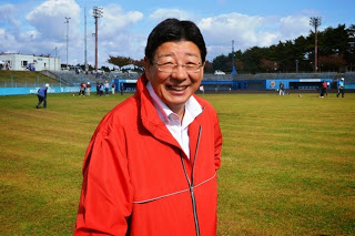 大会が行われている広いグラウンドで、赤いジャンパーを着た佐藤町長がカメラに向かって笑顔を見せている様子をアップで撮影した写真