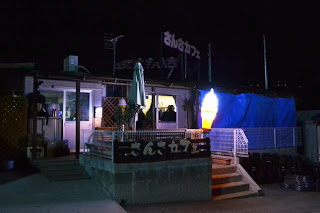 夜間に屋外の建物のテラスに明るく光る大きなアイスクリームの形をした看板があり、テラスの前に手すりや階段があるさんカフェの外観を写した写真