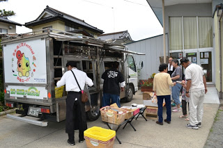 コミュニティセンター前に移動販売車が停車しており、いろいろな品物がテーブルの前にも並べられ、買い物に来た住民の方が品物を選んでいる様子の写真