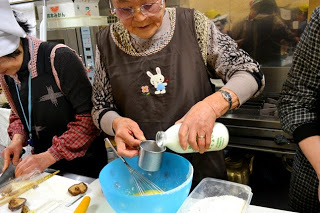 ウサギの刺しゅうがついたエプロンを着た高齢の女性が、大きなボウルに牛乳瓶のような容器から液体を注ぎながら料理をしている調理中の写真