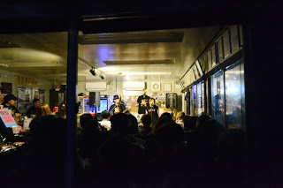 外からガラス越しにさんさカフェの室内を見た視点で、室内には多くの人が集まり、イベントを楽しんでいる様子の写真