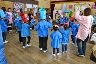 青いスモックを着た園児たちが赤や青の大きな手作りの鬼のお面をかぶって輪になって踊っている室内の写真