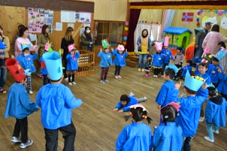 青いスモックを着た園児たちが赤や青の大きな手作りの鬼のお面をかぶって輪になり、その中央で寝転ぶ子供、周りに保護者やピンクのエプロン姿の保育士がカメラ撮影や見守りをしている園内の写真