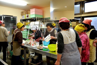 調理室で10人ほどの女性たちがエプロンと三角巾をつけて、ボウルに材料を入れて作業をしている女性の動きを参加者が見ている様子の写真