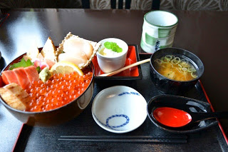 いくらや刺身がのった豪華な海鮮丼に、味噌汁、茶碗蒸し、湯呑が付いた和食の定食が黒いテーブルの上に並んでいる写真