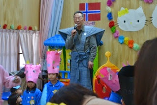 甲冑風の羽織付きの着物を着た男性がマイクで話をし、前列にピンクの鬼のお面風の帽子をかぶった園児たちが並んでいる様子を写した写真