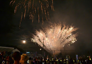 夜の会場で観客たちが見上げる中、空と地上から複数の花火が一斉に打ち上がっている迫力ある花火大会の様子の写真