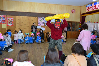 青いスモックを着た子どもたちが座っていて中央に赤鬼の面をかぶり、黄色い金棒の鬼役の男性が立っている節分の日の写真