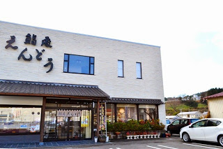 白い車が停まっており、中央に和風の屋根や鉢植えが並ぶ入口がある、「鮨処えんどう」と書かれた白い外壁の建物外観の写真