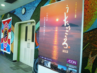 会場の出入口ドアの隣に、太陽の光で海辺の空が赤く染まる風景の大きなポスターが貼られていて、ポスターには「うたごころ 2012」と書かれている屋内の一角の写真