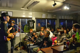 室内で観客が笑顔で音楽ライブを楽しみ、ギター演奏をする男性とマイクで話す男性がステージに立っている様子の写真
