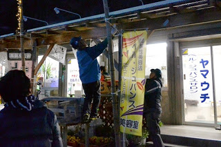 商店街の店先で、作業着を着た人が脚立に登って柱にイルミネーションを設置している写真