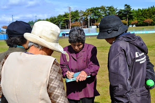 芝生の上で女性がスコアを書き込んでおり、周囲に他の参加者が集まって見ている様子の写真