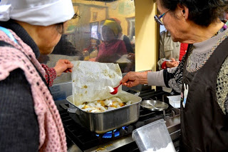 ガスコンロの上で女性が紙の落し蓋をかぶせた鍋の中の料理に、砂糖の様な白い粉をふりかけている様子が写っている煮込み料理の調理中の写真