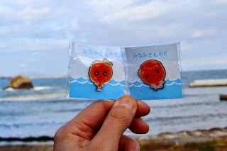 海を背景に「みなみさんりく」と書かれたパッケージの中に、いくらのキャラクター「イクランとキララン」が描かれた可愛らしいピンバッジを持っている写真