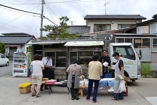 移動販売車の前に数人の住民の方が訪れ、品物を見ている様子を写した写真