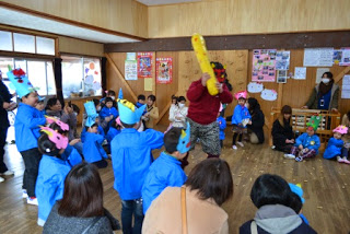 中央の赤鬼の面をかぶり、黄色い金棒の鬼役の男性にむかって青いスモックの園児たちが手には紙で作られた豆の入れ物や豆を持って鬼に向かって「豆まき」をしている様子を写した写真
